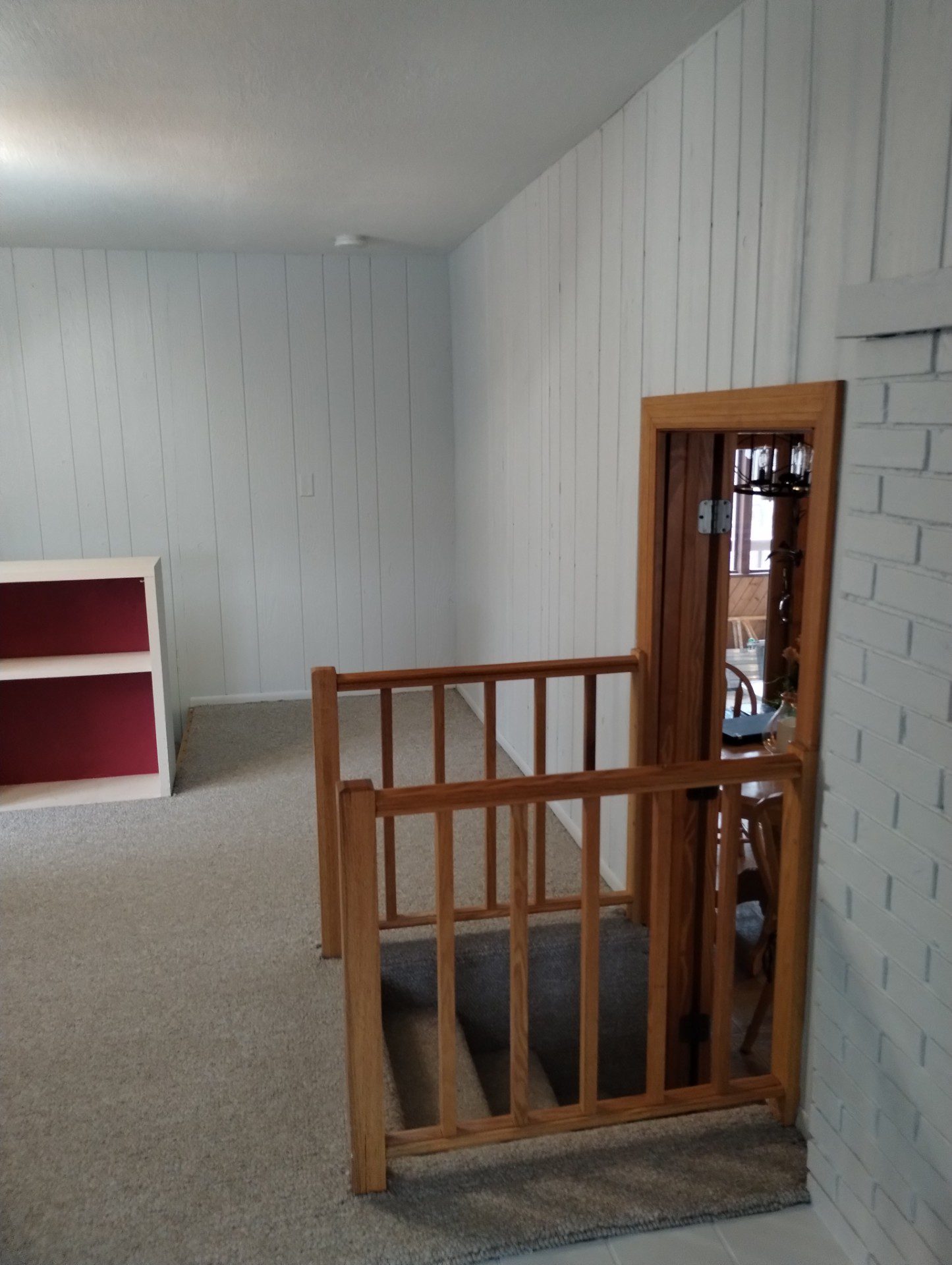 Freshly painted upstairs landing and hallway with light gray painted vertical wood panel walls, white painted brick fireplace, beige carpet, oak stair railing and doorway trim, and a built-in white shelving unit with a red interior